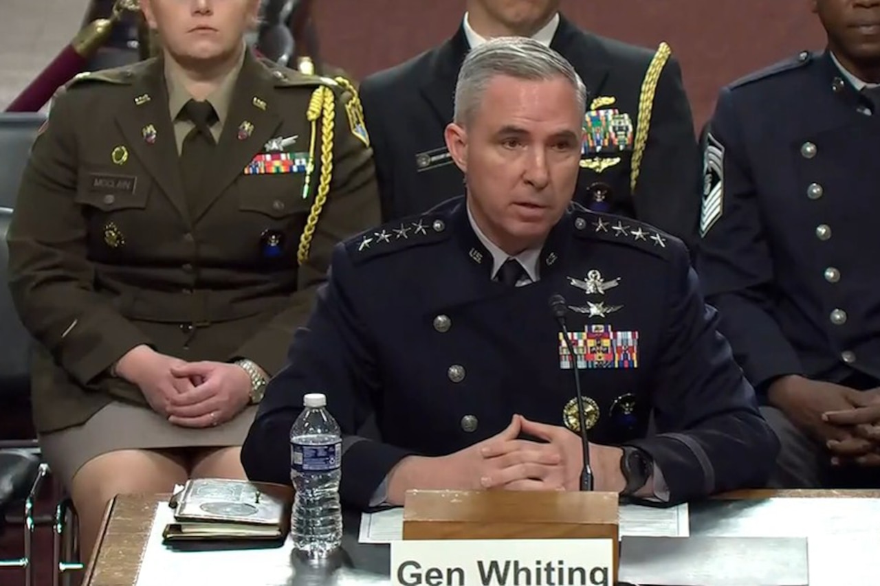 A man wearing a formal military uniform speaks into a microphone while seated at a table. There are three people in similar attire seated behind him.