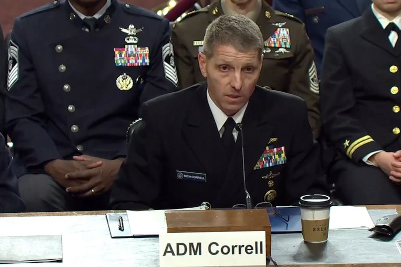 A man wearing a formal military uniform speaks into a microphone while seated at a table. There are three people in similar attire seated behind him.