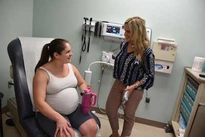 Pregnant woman sits in a doctor's office chair while speaking to a standing woman