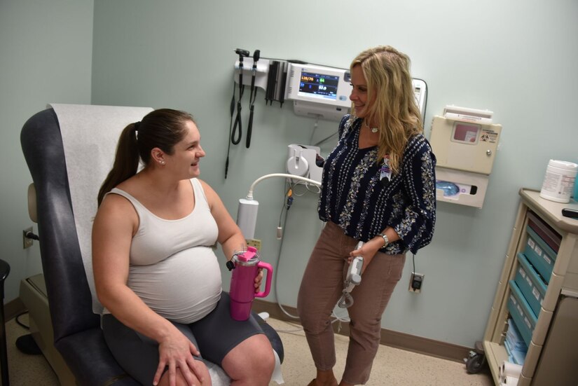 Pregnant woman sits in a doctor's office chair while speaking to a standing woman