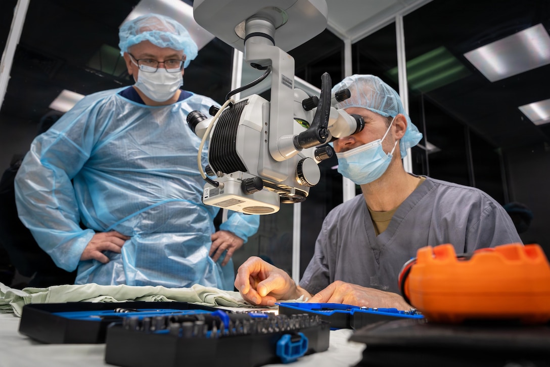 A doctor looks through a microscope while sitting at a table as a fellow doctor wearing scrubs watches.