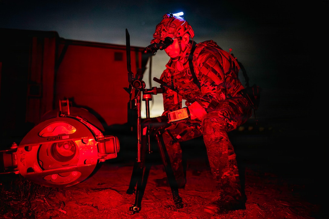 A sailor in tactical gear sets up an X-ray in a field in the dark illuminated by red light.