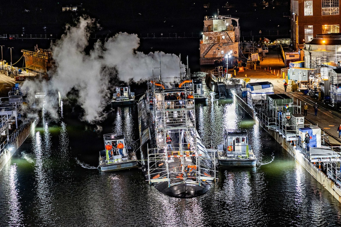 Smoke blows in the air as a submarine enters a dock in the dark illuminated by street and building lights.