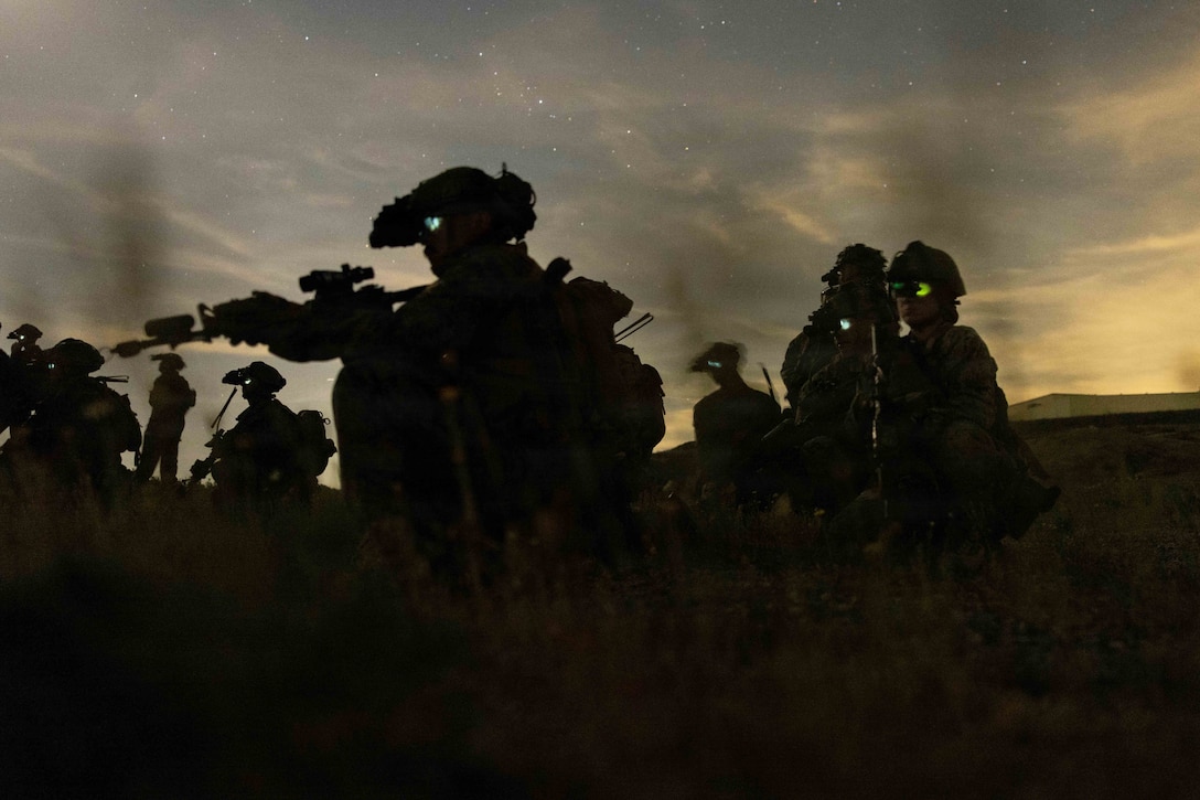 Marines in tactical gear move through a field while aiming weapons under a starry night sky illuminated by a yellow glow.