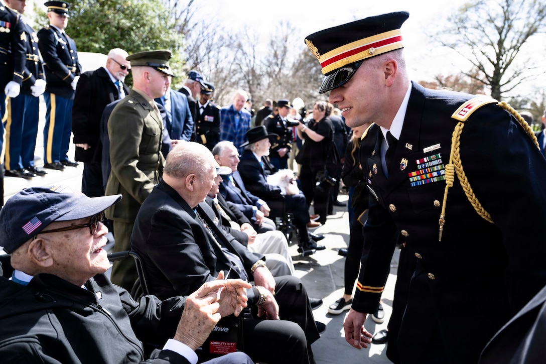 A service member in ceremonial dress speaks to a seated veteran during a ceremony.