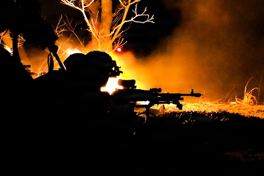A soldier lies on the ground with a weapon as an orange fireball sits to the left near a tree as seen in silhouette.