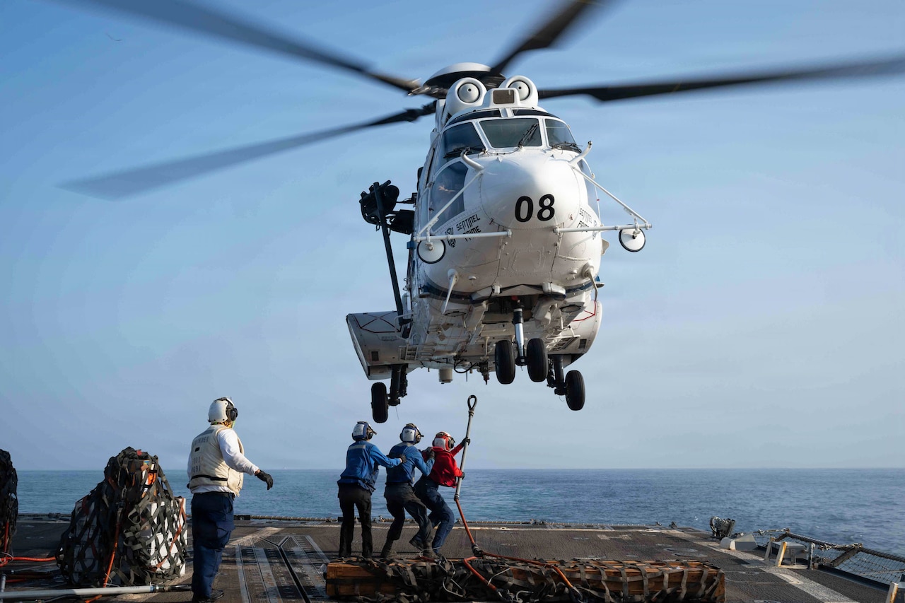 Three sailors hold one another in a line as the leader extends a hook toward a hovering helicopter aboard a ship at sea during the day.