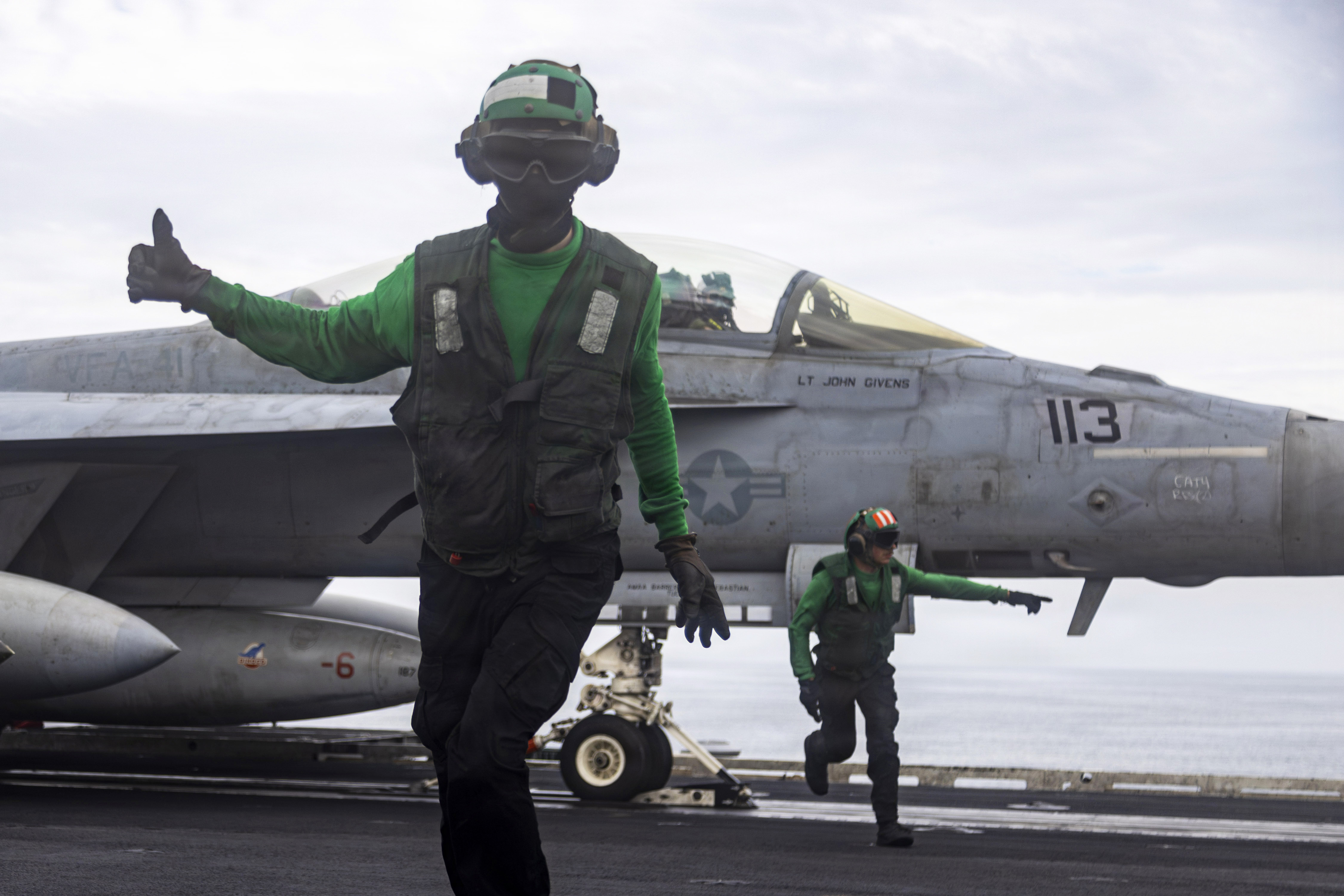 Two sailors wearing green vests and helmets give signals while moving away from an aircraft aboard a ship at sea.