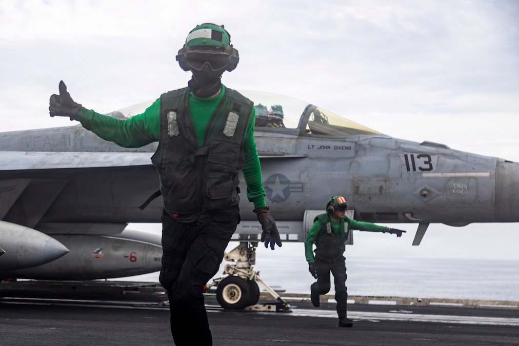 Two sailors wearing green vests and helmets give signals while moving away from an aircraft aboard a ship at sea.