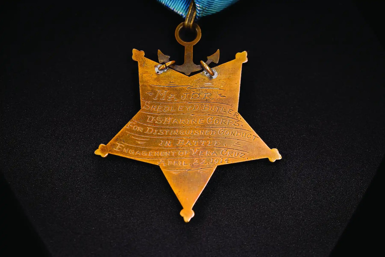A gold-colored, five-pointed star with an etched inscription hangs from a gold-colored anchor, hanging from a ribbon.
