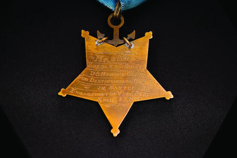 A gold-colored, five-pointed star with an etched inscription hangs from a gold-colored anchor, hanging from a ribbon.