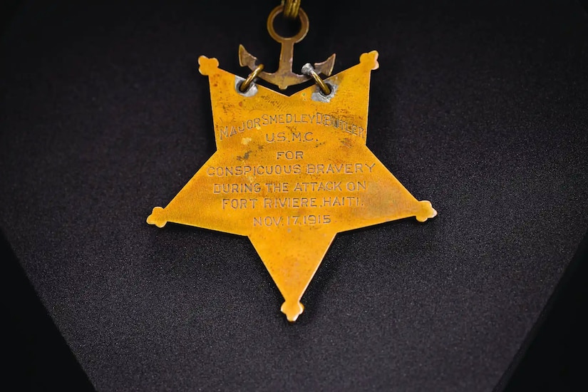 A gold-colored, five-pointed star with an etched inscription hangs from a gold-colored anchor, hanging from a ribbon.