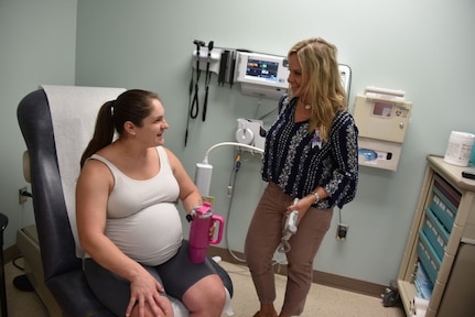 Pregnant woman sits in a doctor's office chair while speaking to a standing woman