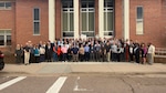 A large group of people pose for a photo in front of a brick building.