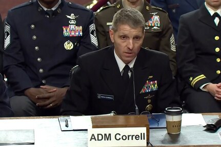 A man wearing a formal military uniform speaks into a microphone while seated at a table. There are three people in similar attire seated behind him.