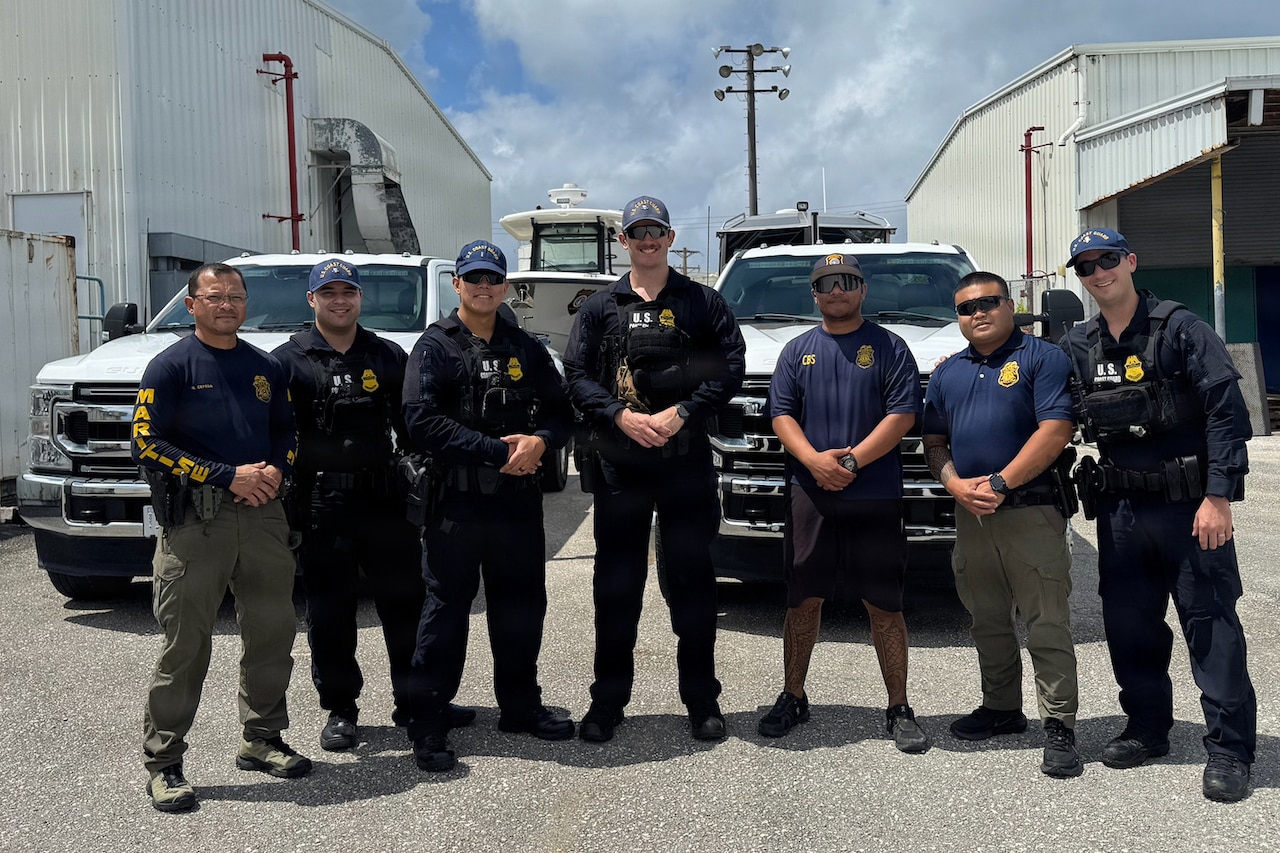 Four men wearing military uniforms and three men dressed in law enforcement attire pose for a photo in front of two trucks and a small boat on a trailer.