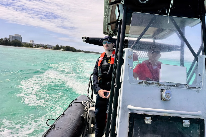 A man wearing a military uniform and life jacket looks out at the water from a small boat while another man drives the boat.