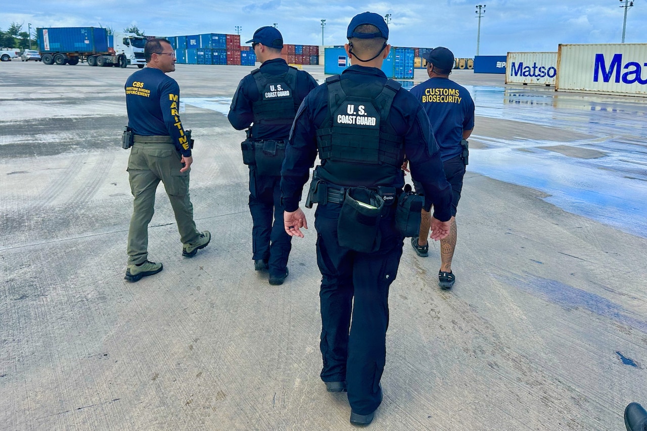 Two men wearing military uniforms and two men dressed in law enforcement attire walk together around a port.