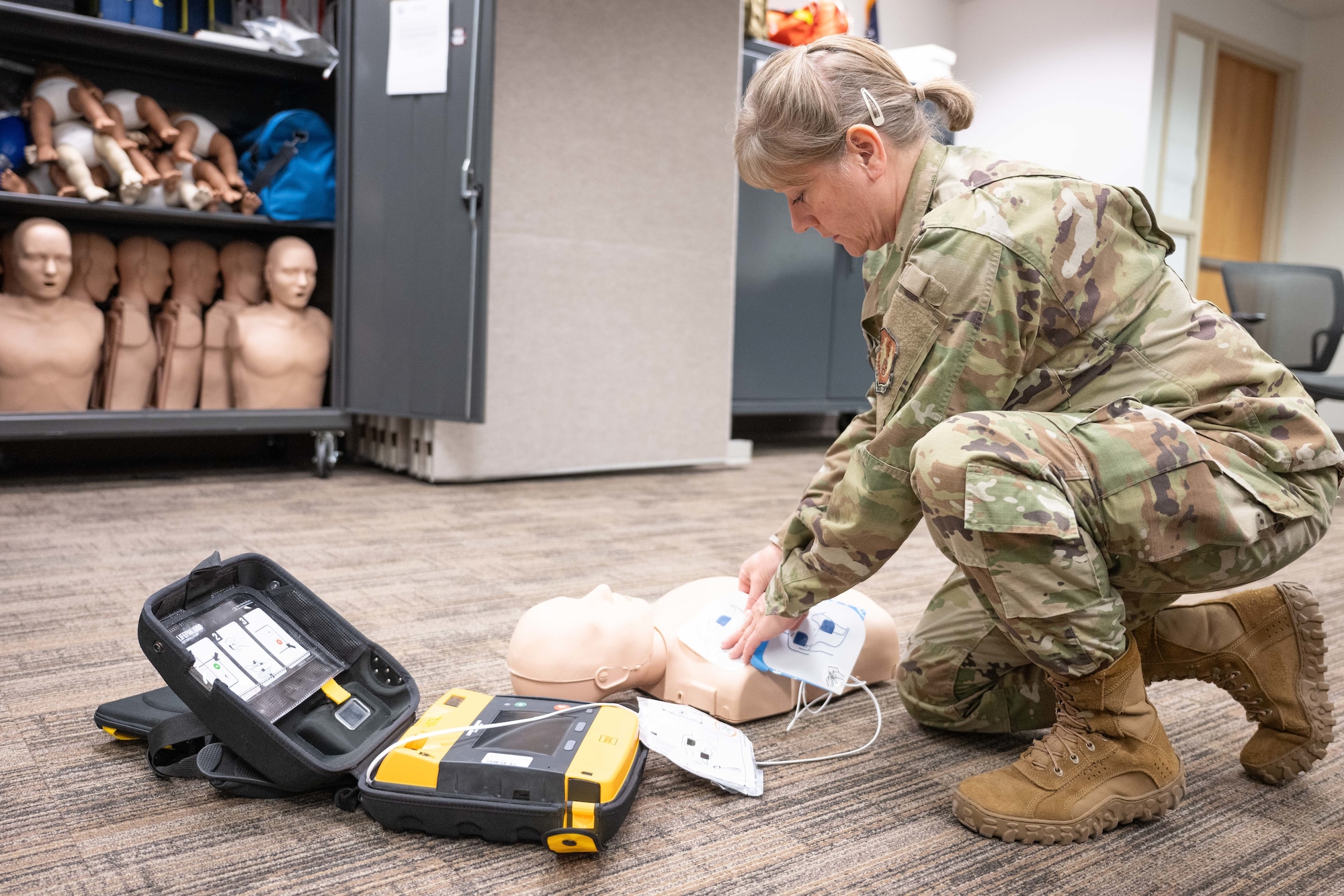 A person in military uniform practices using an automated external defibrillator on a training manikin in a classroom setting.