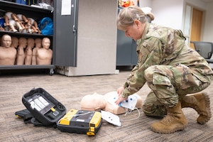 A person in military uniform practices using an automated external defibrillator on a training manikin in a classroom setting.