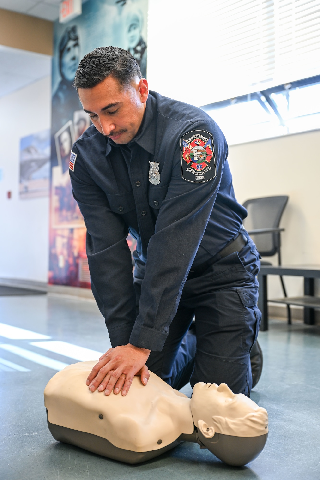 A firefighter in uniform performs chest compressions on a CPR training manikin in a brightly lit room.