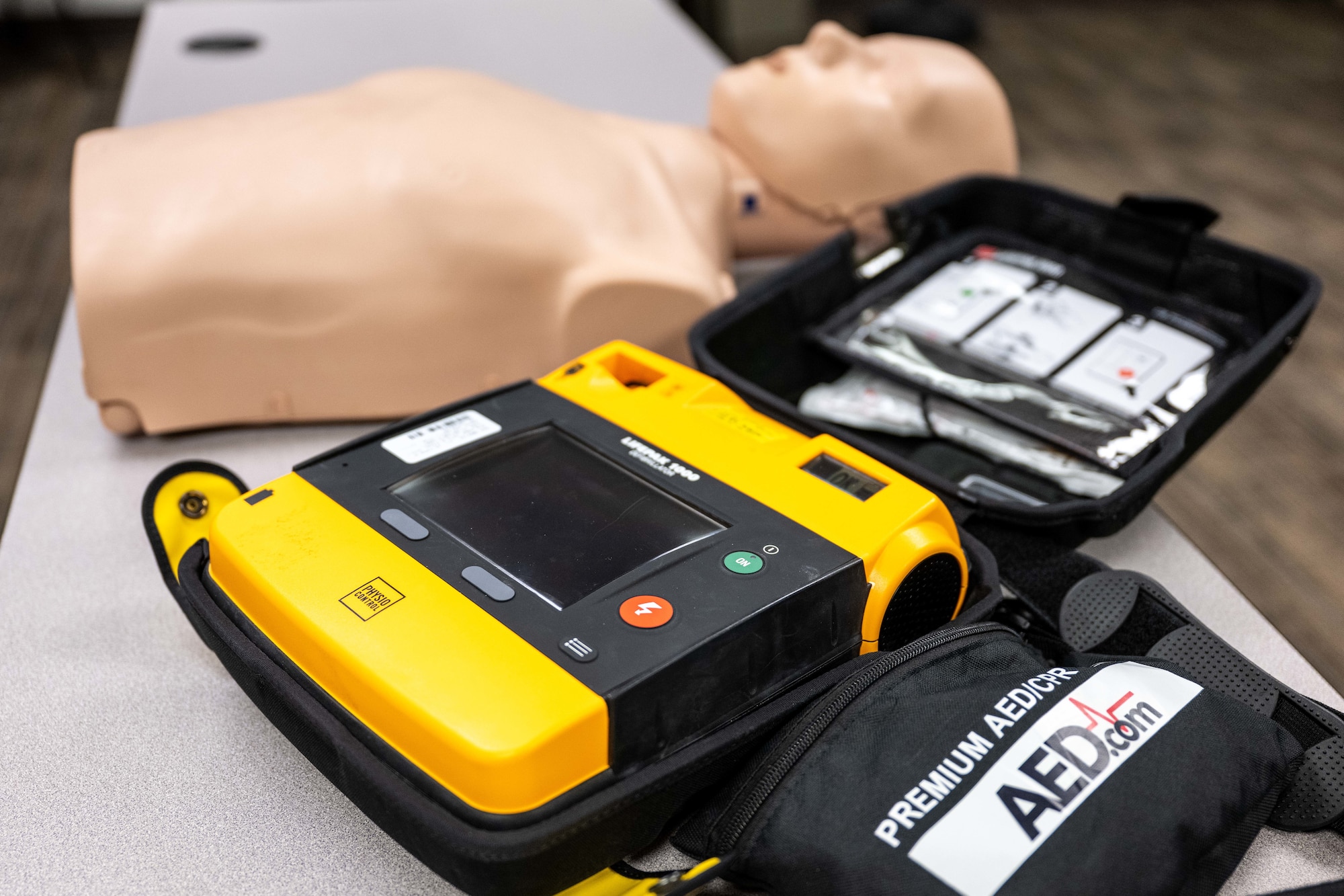 An Automated External Defibrillator in its case sits next to a CPR training manikin.