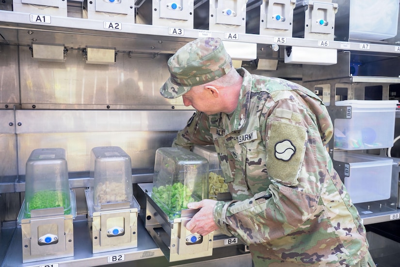 A soldier wearing a camouflage military uniform loads a container of vegetables into an outdoor, mobile military container kitchen.