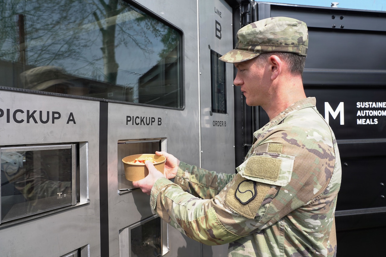 A soldier wearing a camouflage military uniform retrieves a bowl of food from an outdoor military container kitchen.