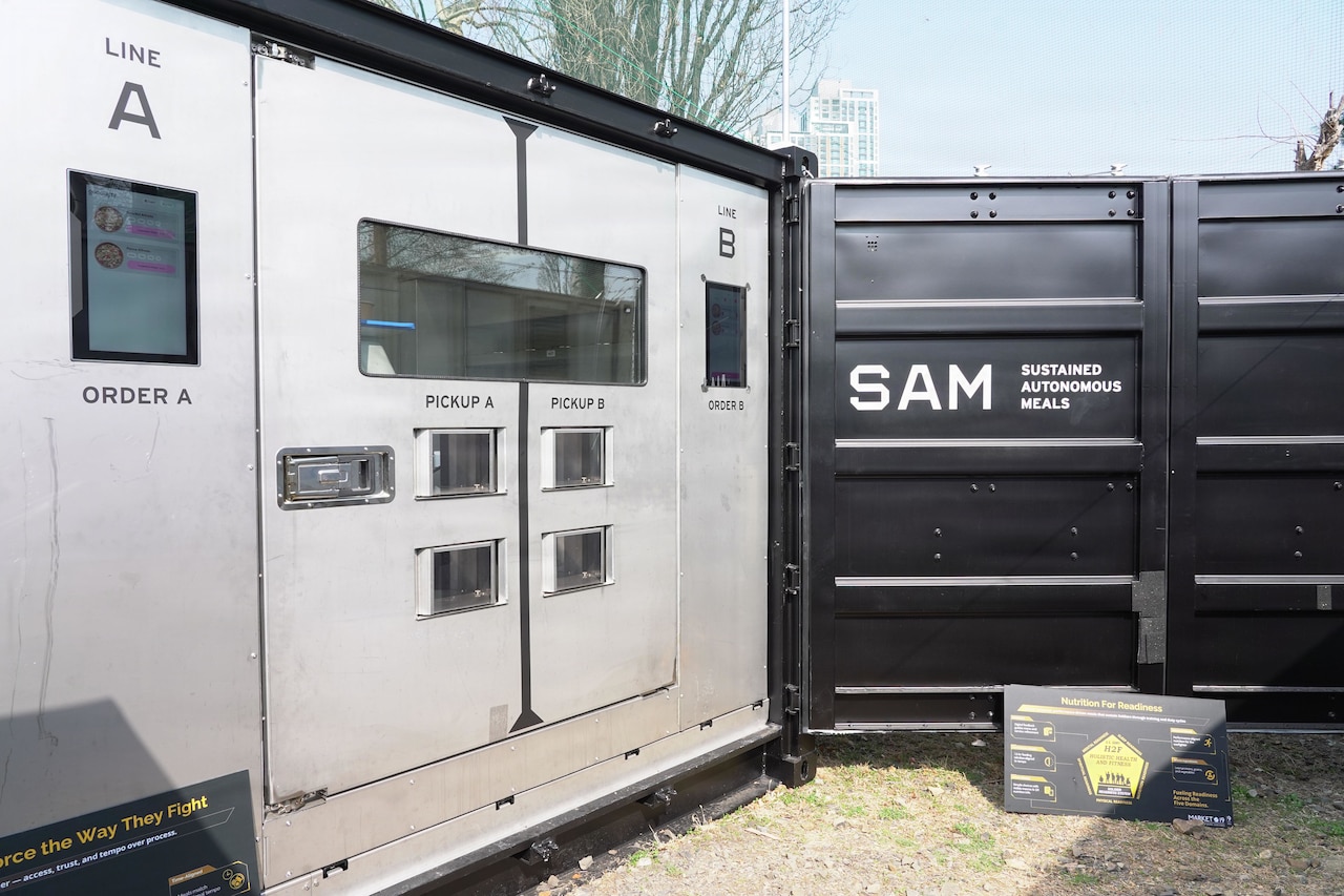 Several large containers sit outside in a gravel area. Each is labeled with kitchen words such as “line,” “order” and “pickup;” the container on the right is labeled “SAM, sustained autonomous meals.”