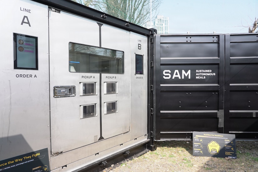 Several large containers sit outside in a gravel area. Each is labeled with kitchen words such as “line,” “order” and “pickup;” the container on the right is labeled “SAM, sustained autonomous meals.”