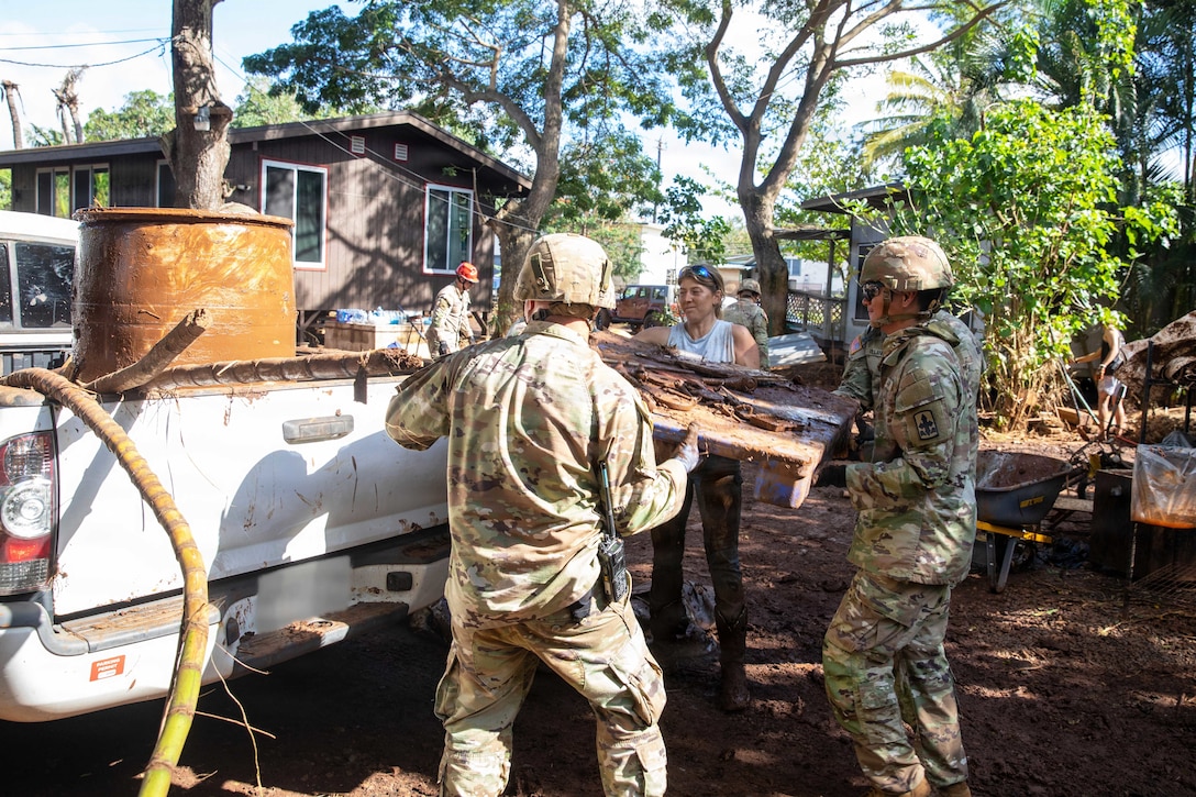 Soldiers and a civilian lift debris into a pickup truck in a shaded, wooded area near a brown house.