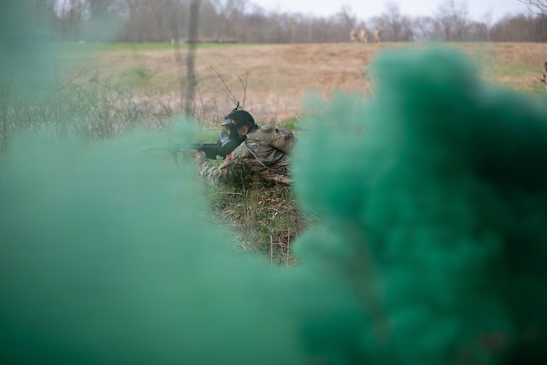 A soldier wearing a protective mask lies in the grass while aiming a weapon as green clouds of smoke fill the foreground.