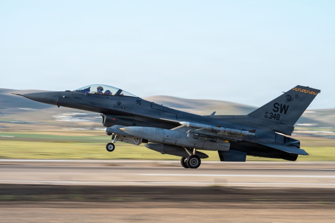 A blurred image of an aircraft landing on a tarmac with mountains in the background during the day.