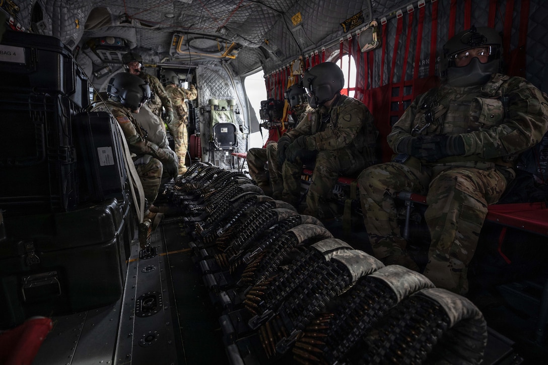 Soldiers wearing helmets sit and stand aboard a dark military aircraft.