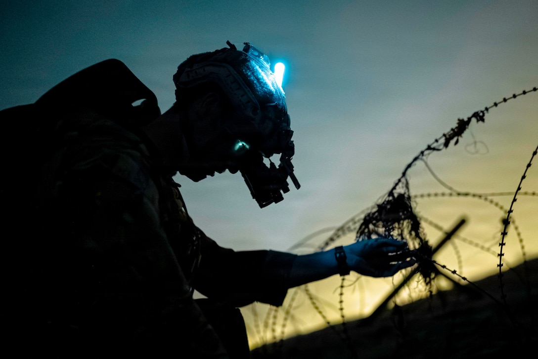 A sailor wearing a helmet and night vision lens prepares to cut barbed wire in the dark illuminated by a headlight and yellow glow.