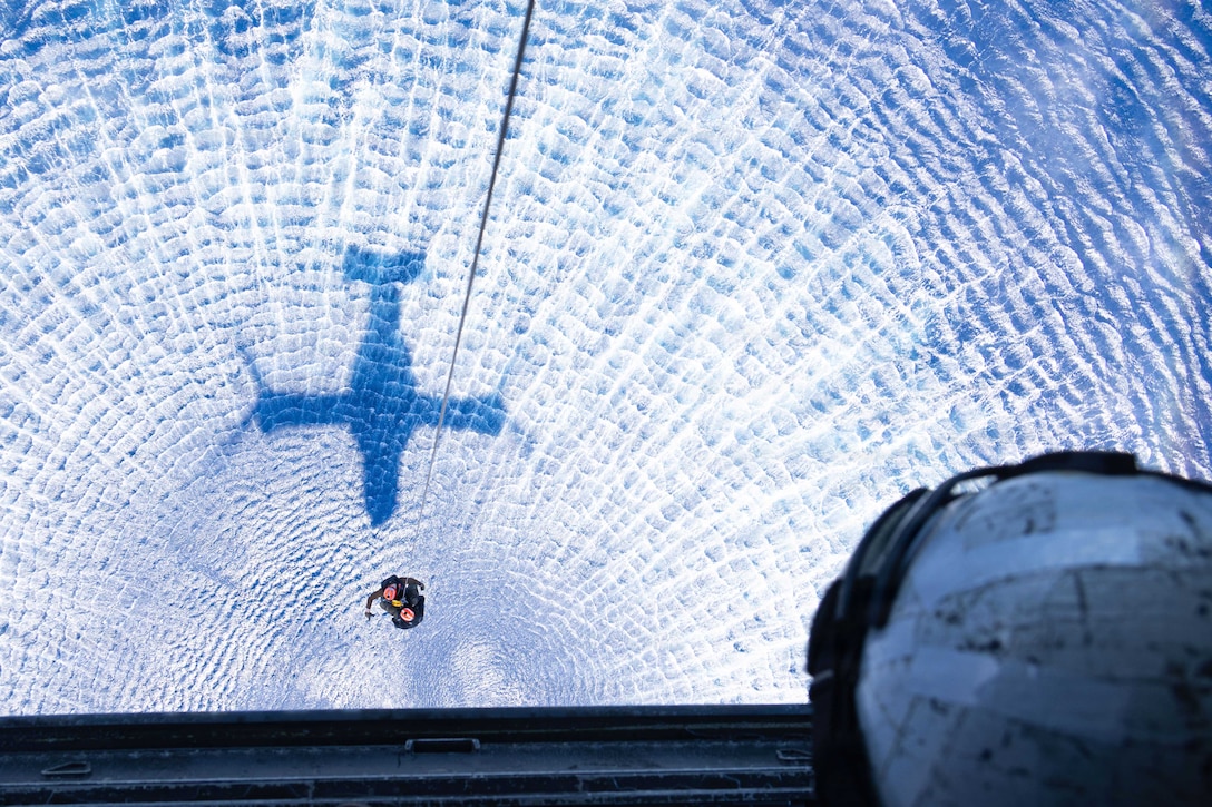 A sailor watches as fellow sailors are hoisted toward an airborne aircraft from a body of wavy waters near the shadow of the aircraft.
