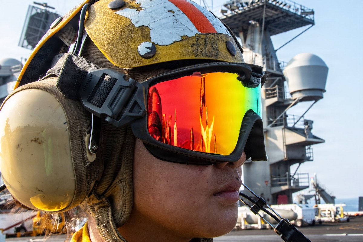 A close-up of a sailor wearing a helmet, earmuffs and reflective goggles aboard a ship at sea.