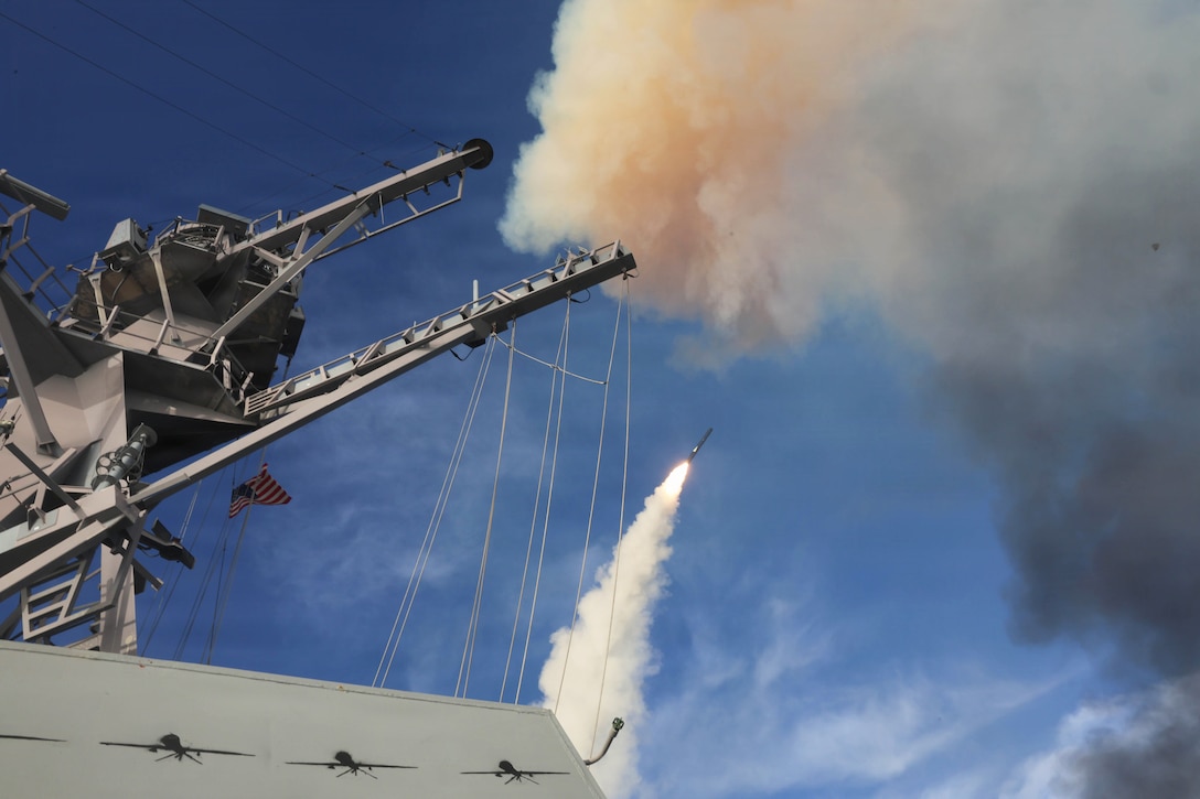 A side view of a ship firing a missile into a blue sky, leaving a trail of smoke.