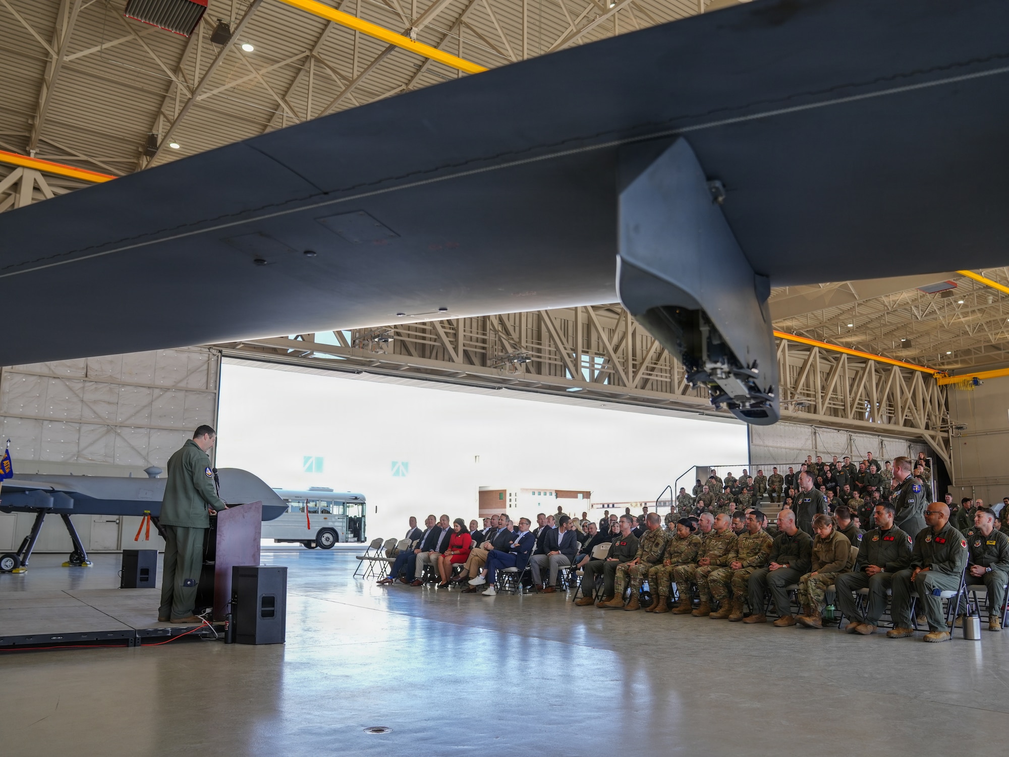 A large crowd of U.S. Air Forces members are sitting and standing on bleachers while looking at a U.S. Air Force member speak from a podium in an aircraft hangar.