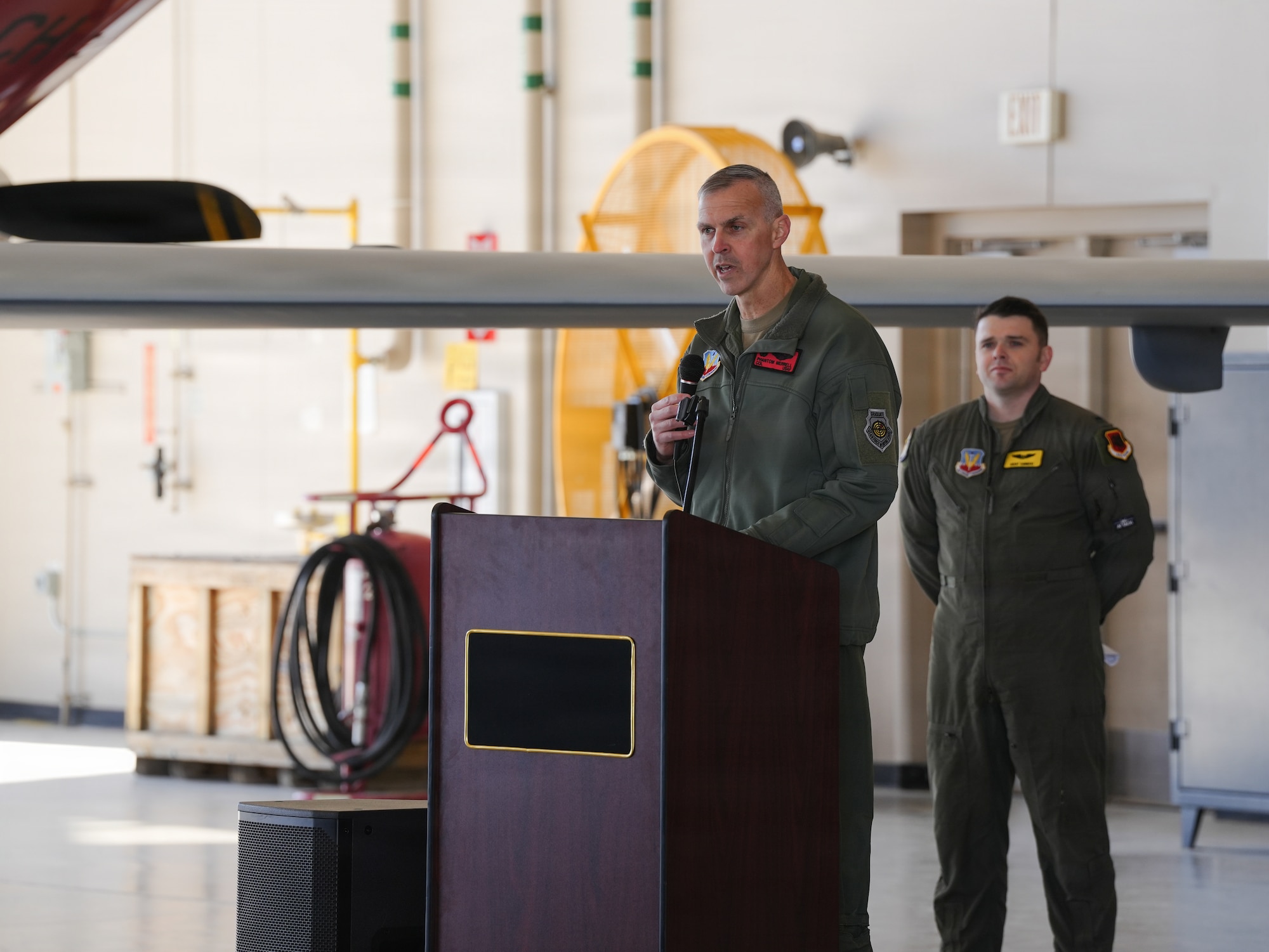 A U.S. Air Force Col. delivers remarks from a podium with a microphone in an aircraft hangar.