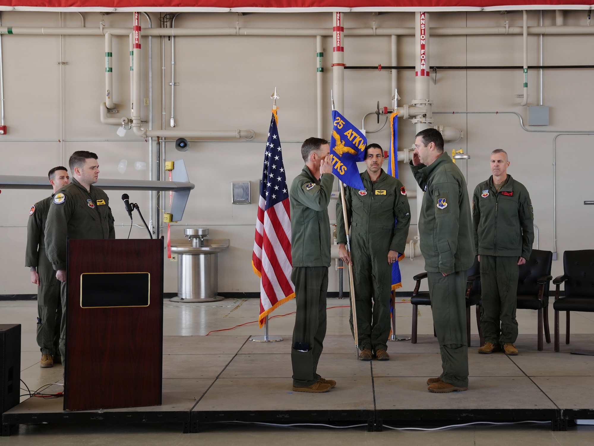 Two U.S. Air Force officers are facing each other saluting on a stage in an aircraft hangar.
