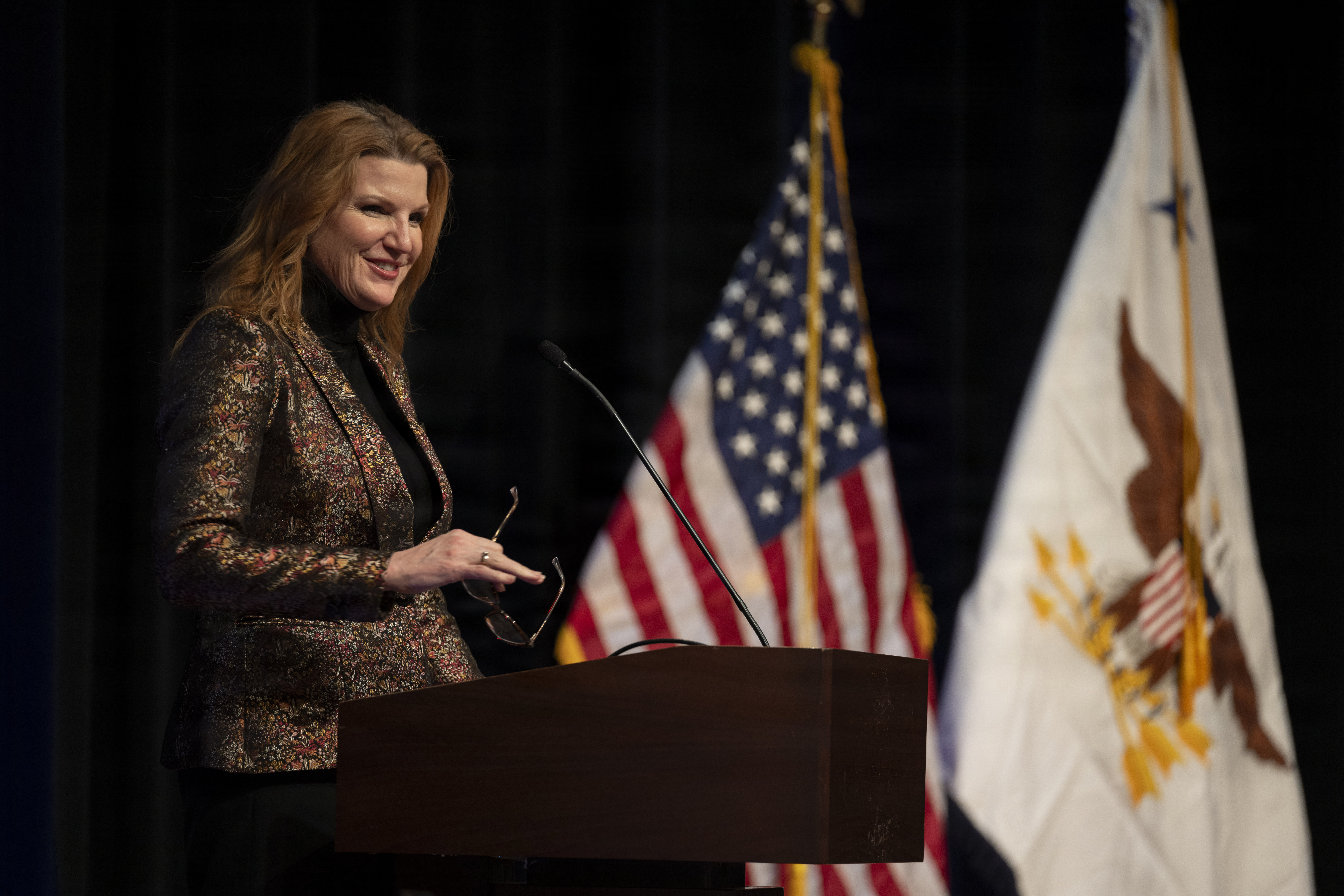 A woman in business attire stands behind a lectern while holding a pair of glasses and smiling. Behind her are an American flag and a flag with an eagle.