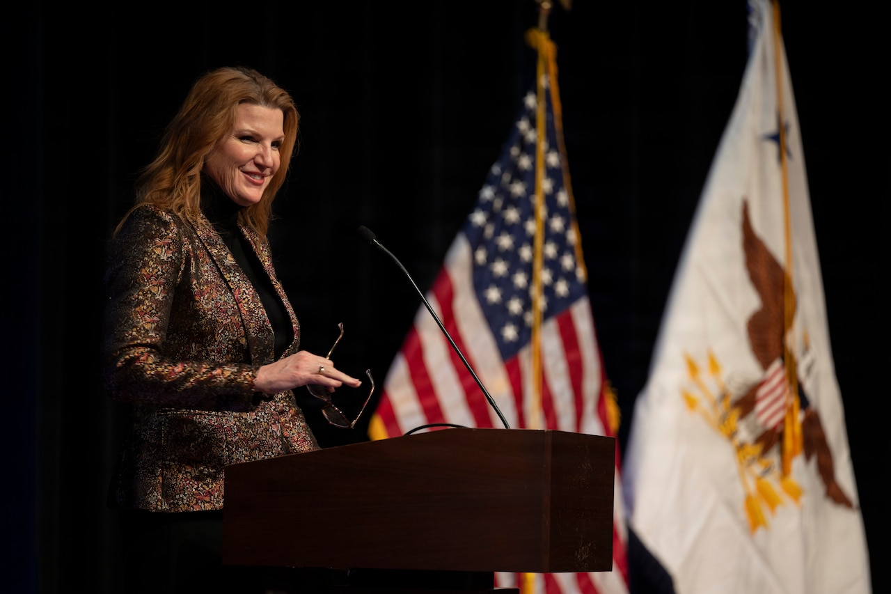 A woman in business attire stands behind a lectern while holding a pair of glasses and smiling at something off-screen. Behind her are an American flag and a flag with an eagle carrying three arrows in the center.
