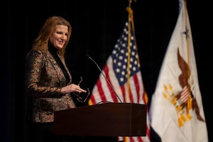 A woman in business attire stands behind a lectern while holding a pair of glasses and smiling at something off-screen. Behind her are an American flag and a flag with an eagle carrying three arrows in the center.