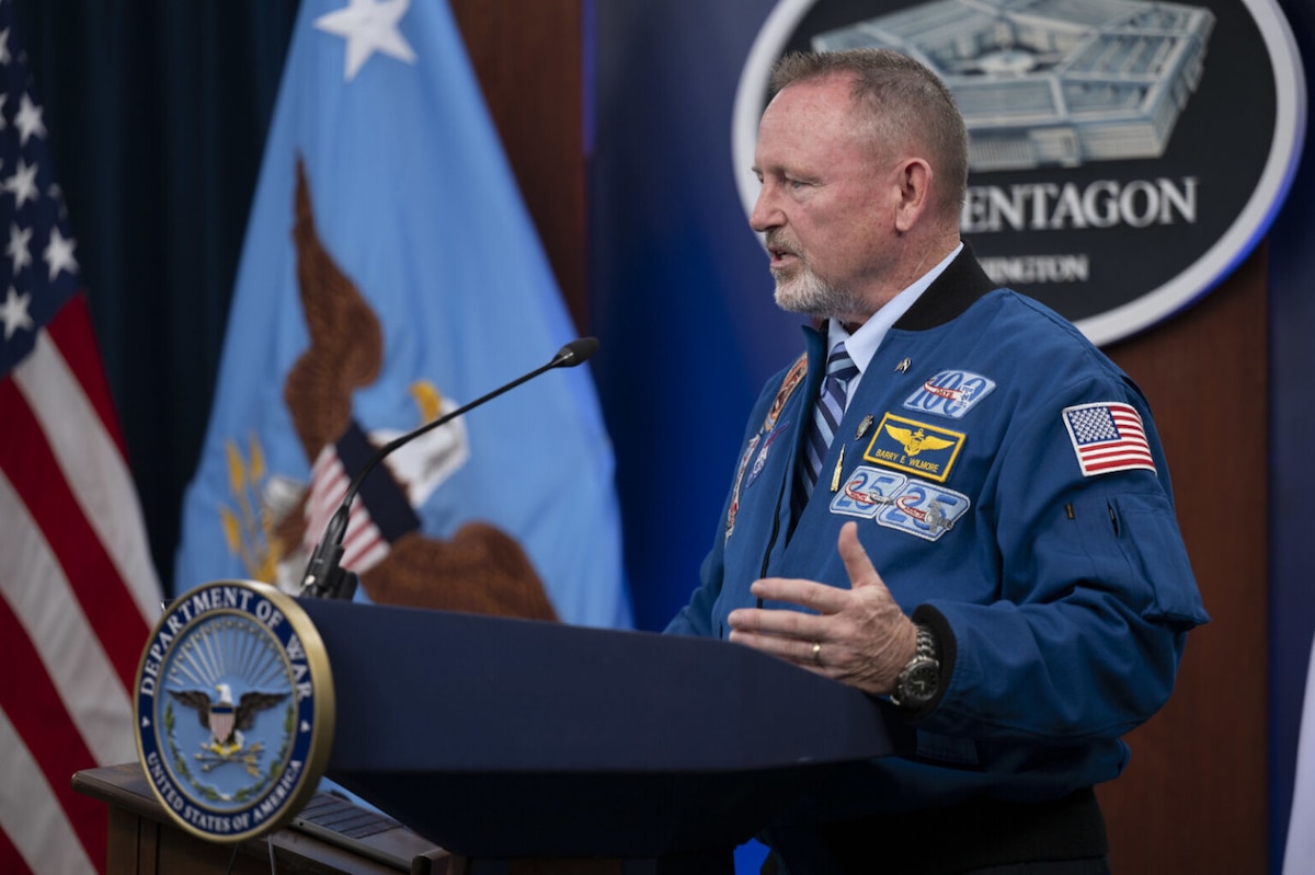 A man wearing a blue jacket with multiple patches stands behind a lectern and speaks with a sign behind him that reads, "The Pentagon."