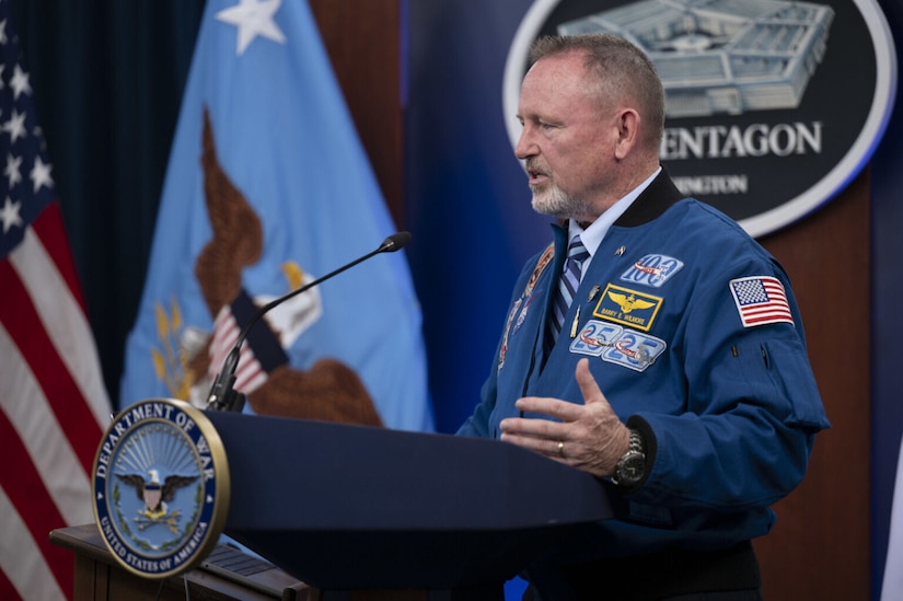 A man wearing a blue jacket with multiple patches stands behind a lectern and speaks with a sign behind him that reads, "The Pentagon."