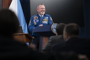 A man wearing a blue jacket with multiple patches stands behind a lectern and speaks to an audience.