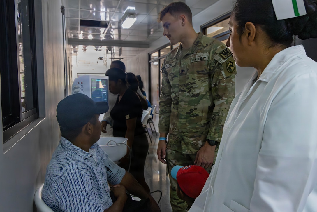 A military member and medical personnel in a white lab coat monitor a patient's vitals
