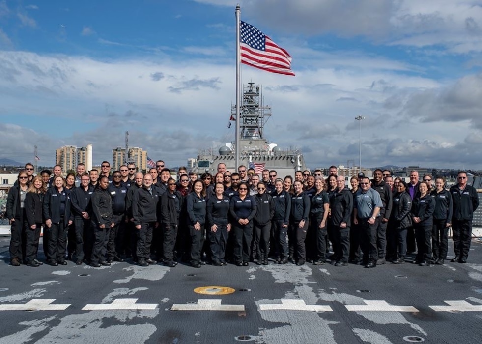 Members pose on the deck of a naval ship. There is an American flag flying in the background above the group.