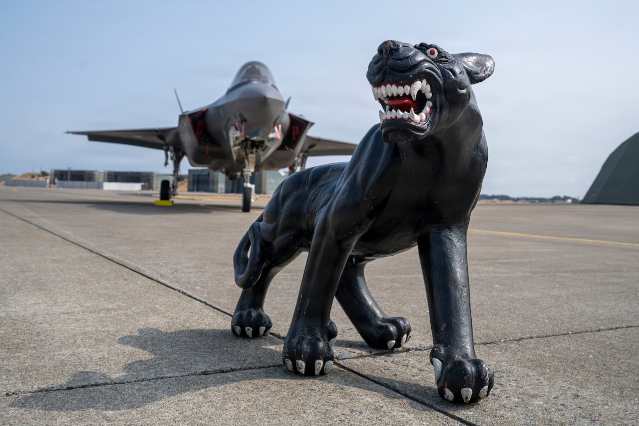 A statue of a panther is displayed in front of a military fighter jet on a flight line.